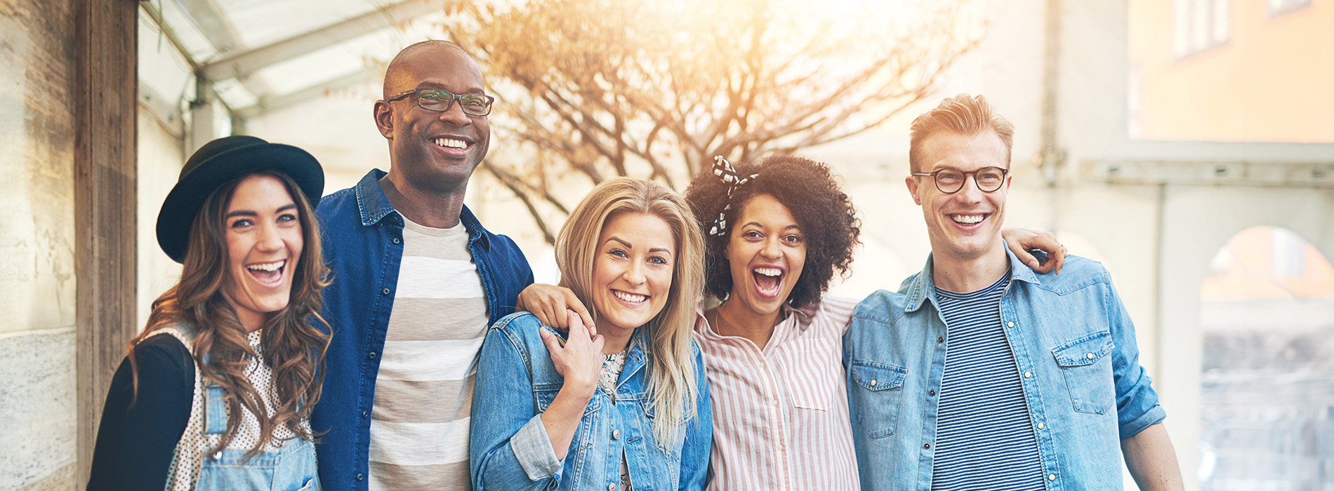 The image shows a group of six people posing together for a photo they are outdoors, wearing casual attire, with a building and trees visible in the background.