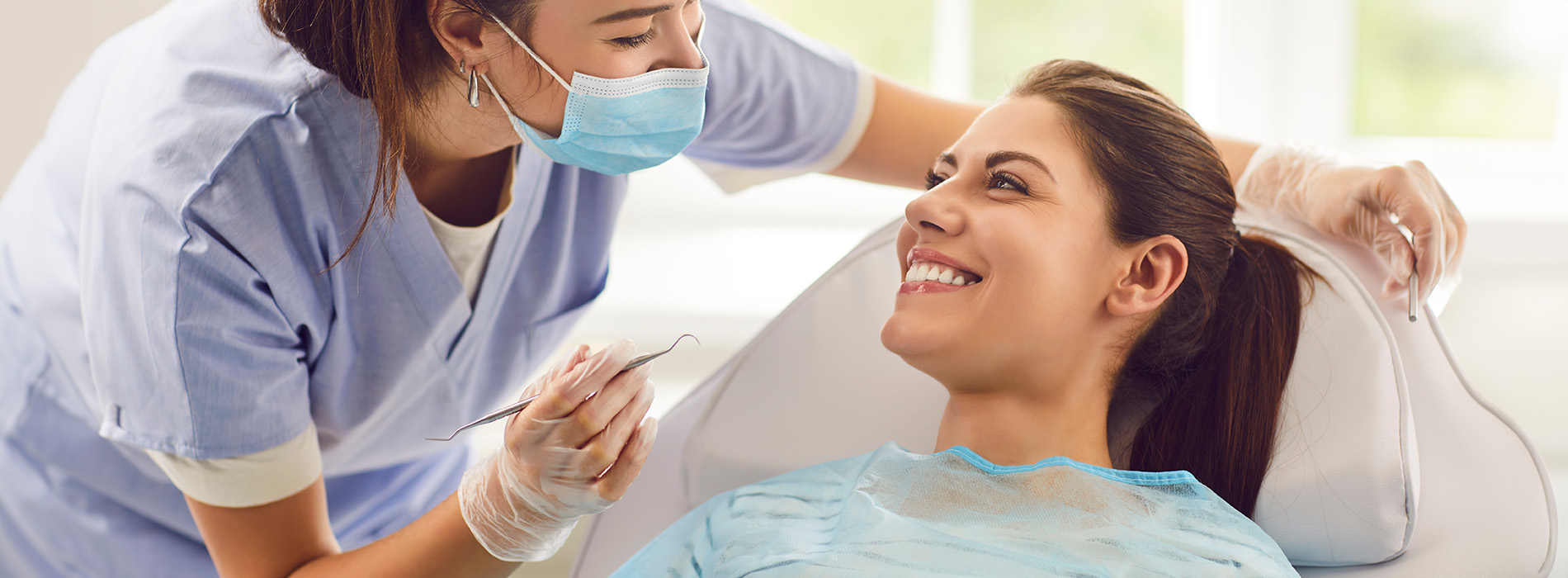 A person is seated in a dental chair, receiving care from a dental professional who stands behind them.