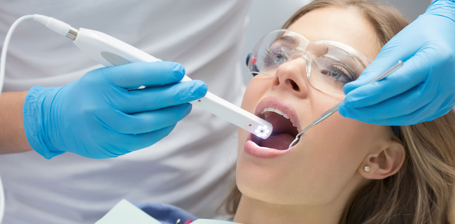 An image of a dental professional using a dental device to clean or examine a person s teeth, with the patient seated in a dental chair and wearing protective eyewear.