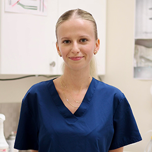 The image shows a person standing in front of a counter with a white background, wearing a blue scrub top and a gold necklace, posing for the camera with a neutral expression.