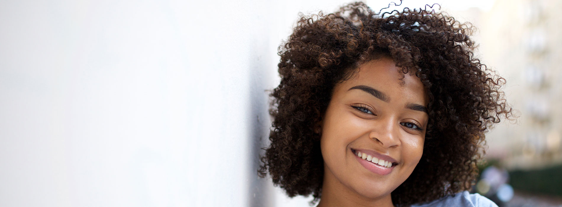 The image is a photograph of a smiling individual with short, curly hair against a blurred background.