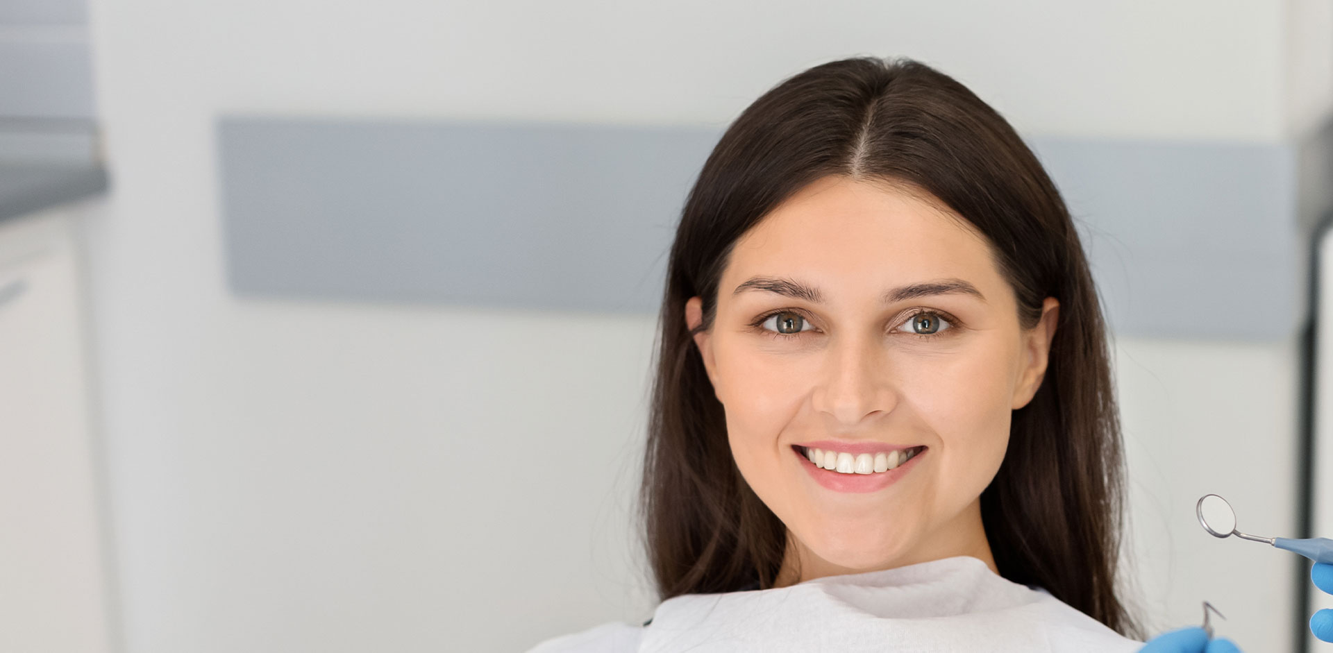 The image shows a woman sitting in a dental chair, smiling at the camera, with a dental practitioner s room visible in the background.