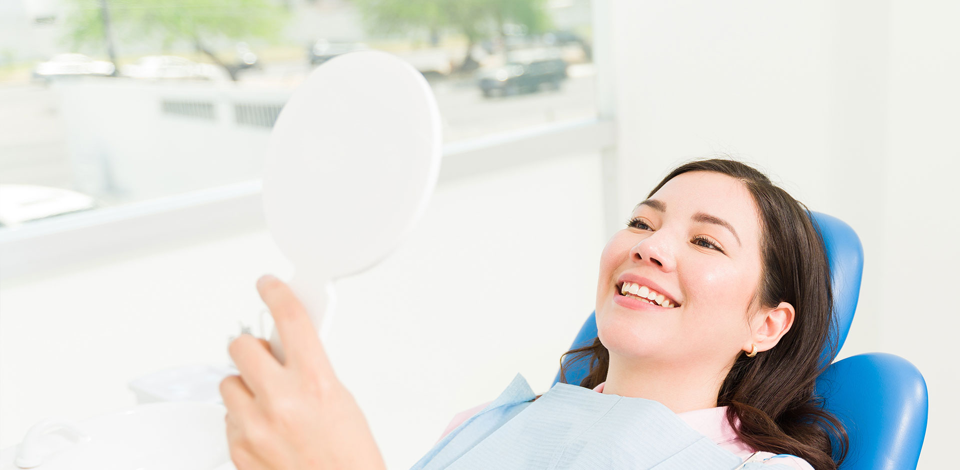 A smiling woman is seated in a dental chair, holding a mirror up to her face while looking at her reflection.