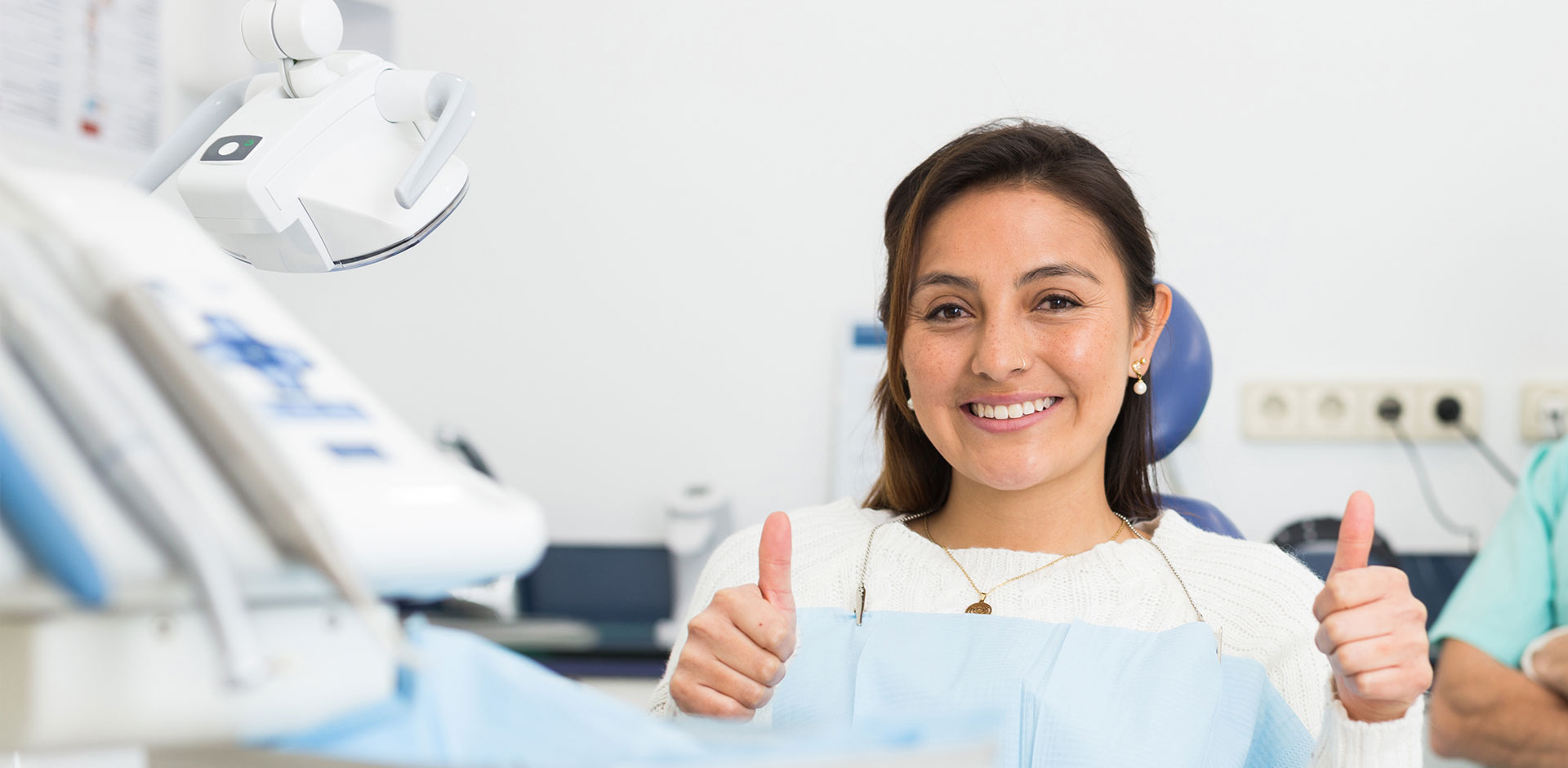 The image shows a person giving a thumbs-up gesture, smiling and wearing a mask, standing in front of medical equipment with a blue background.