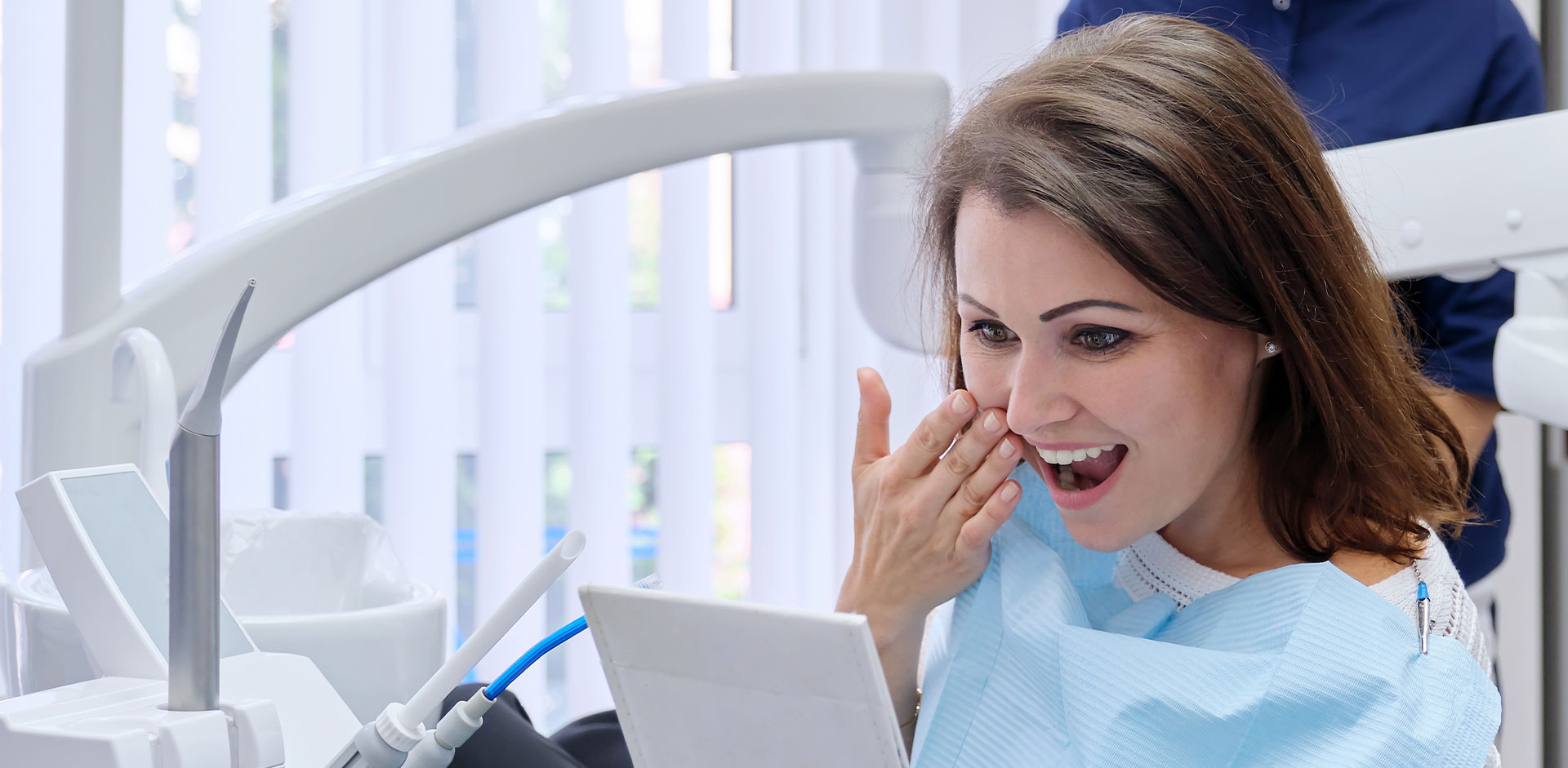 A woman in a dental office, holding her hand to her mouth while seated at a desk with a computer monitor.