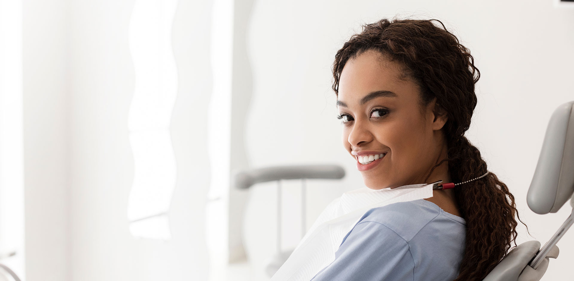 The image shows a smiling woman wearing scrubs, sitting in a dental chair with a dental mirror and light.