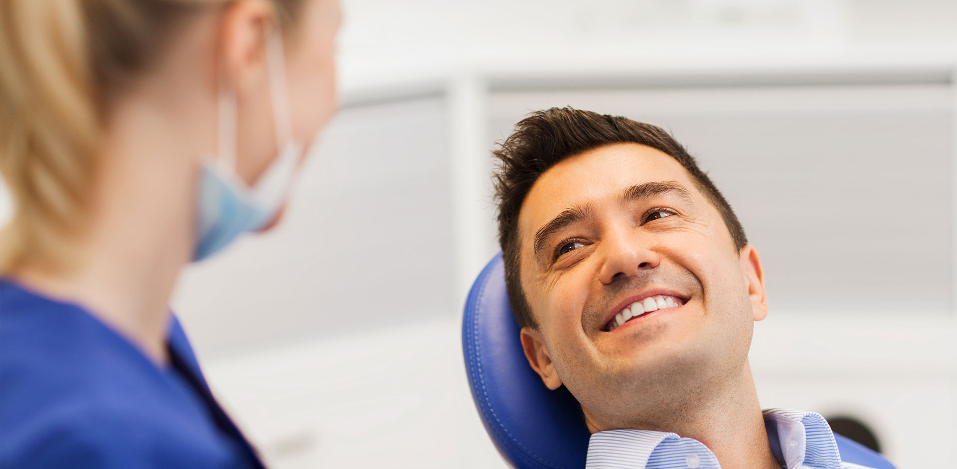 A man in a dental chair is smiling, while a woman in a lab coat stands beside him, also smiling.