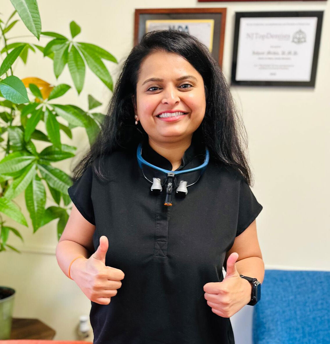 The image shows a person giving a thumbs-up gesture, smiling at the camera. They are wearing a black shirt and have a lanyard around their neck. The individual is standing in an indoor setting with a plant visible behind them.