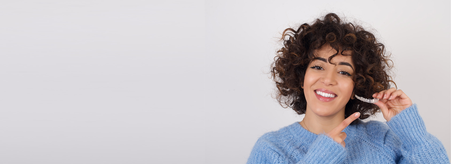 A woman with curly hair is smiling, holding a phone to her ear, and standing against a plain background.