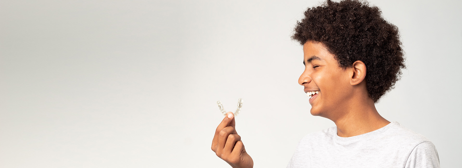 The image features a young person with an open-mouthed smile, holding a flower and looking upwards. They are in front of a plain background.