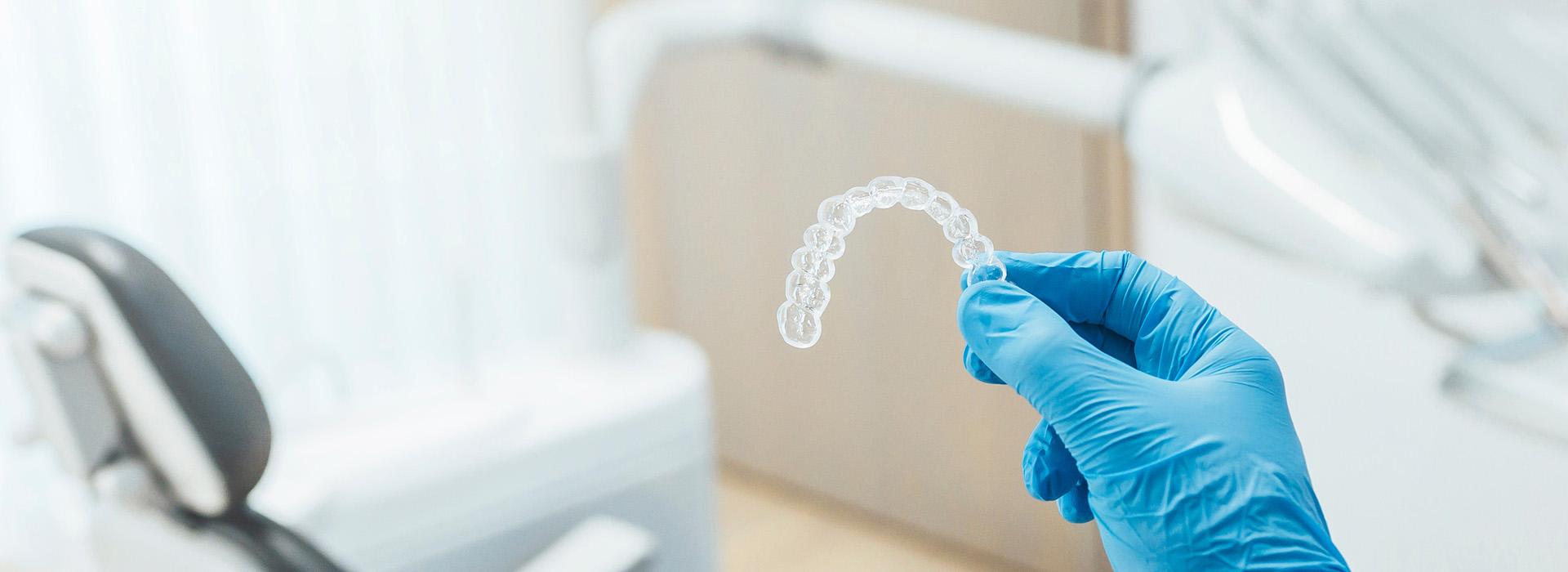 A dental professional wearing gloves is holding a transparent dental impression over a dental chair, with a blurred background showing a clinical setting.