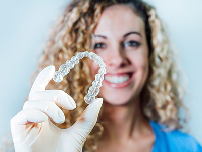 A woman wearing a white glove holds up a clear dental retainer with visible teeth impressions, against a plain background.