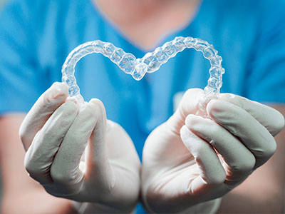 The image shows a pair of transparent, heart-shaped braces being held by a person wearing white gloves and a blue medical uniform.