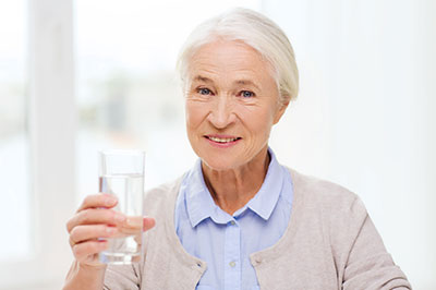 The image shows an elderly woman holding a glass of water and smiling.