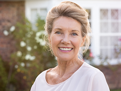 The image shows a woman standing in front of a house, smiling at the camera.