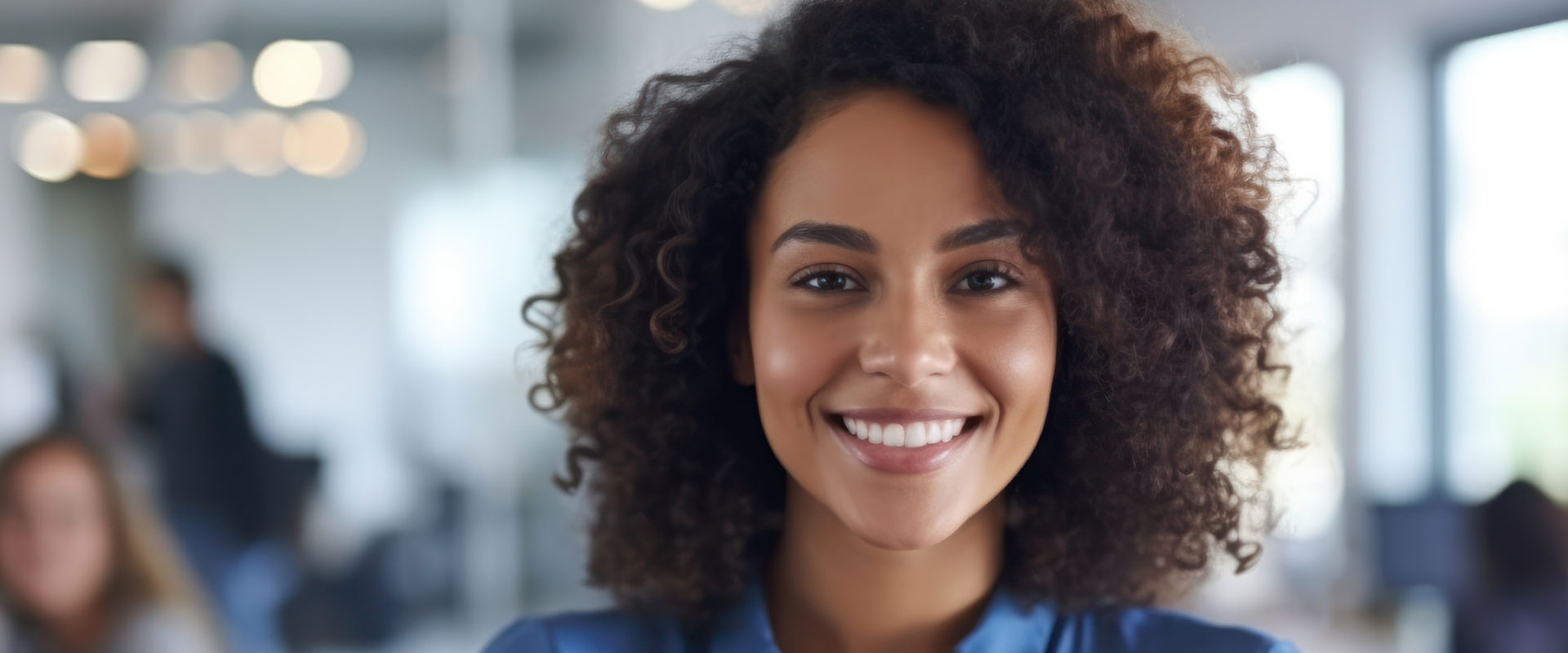 A smiling woman with curly hair, wearing a blue top, in an office environment.