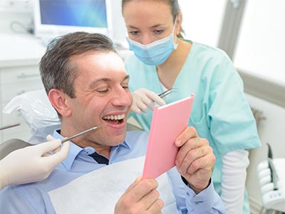 The image depicts a man in a dental chair, holding up a pink card with a smiling expression, while a female dentist looks on, both within a dental office setting.