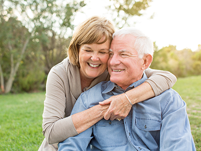 The image shows an elderly couple sharing a warm embrace, with the man wearing a blue shirt and the woman in a brown top. They are seated on grass outdoors during daylight hours.