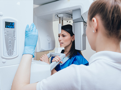 A woman in a blue lab coat is standing next to a large, modern CT scanner machine, with a man wearing gloves looking at the equipment.