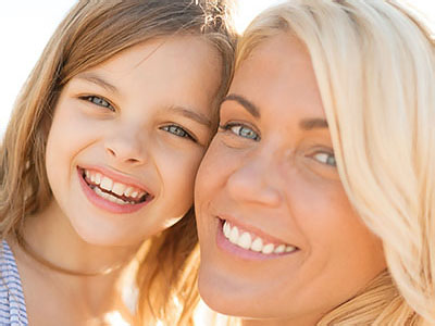 The image shows a woman and a young girl smiling at the camera, with the woman s face partially obscured by the child s head. They appear to be posing for a photo in a sunny outdoor setting.