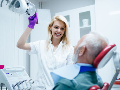 A woman in a white coat is standing next to an older man who is seated and appears to be receiving dental care, with the woman holding a dental mirror.