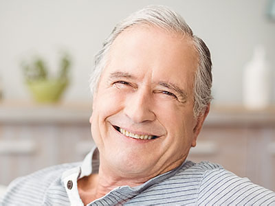 The image shows an elderly man with gray hair, smiling and looking directly at the camera. He is wearing a blue shirt and appears to be in a relaxed setting, possibly a home or office environment.