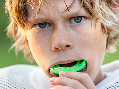 A young boy with blonde hair and a blue eye, wearing a football jersey, is seen blowing a green whistle.